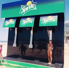 a woman standing in front of a sprite soda machine on the beach with people walking by