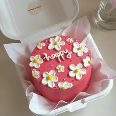 a pink cake with white and yellow flowers on it in a paper box next to a plastic container