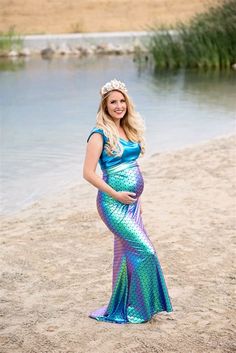 a pregnant woman standing on the beach wearing a blue and green mermaid dress with a tiara