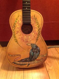 an acoustic guitar painted with a black bird on it's back and sides, sitting on a wooden floor