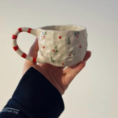 a hand holding a ceramic cup decorated with holly and candy canes, on a white background