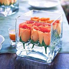 an arrangement of orange flowers in a square vase on a table with plates and silverware