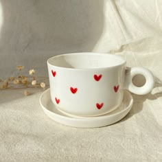 a cup and saucer with red hearts on it sitting on a white cloth next to dried flowers