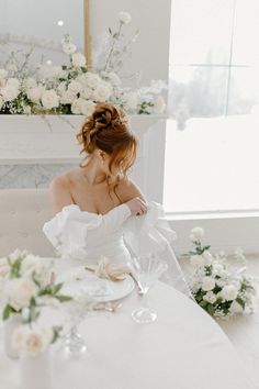 a woman sitting at a table with flowers in front of her, wearing a white dress