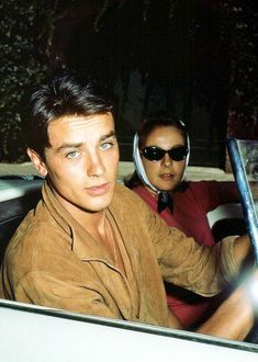 two men sitting in a car, one wearing sunglasses and the other looking at the camera