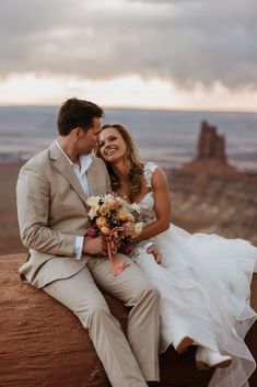 a bride and groom sitting on top of a rock in front of the desert at sunset