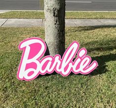 a pink and white sign that says barbie next to a tree