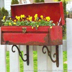 a red box with flowers in it sitting on a white picket fence next to green grass