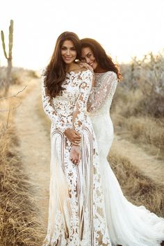 two women in white dresses standing next to each other on a dirt road with cactus trees behind them