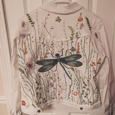 a white jacket with dragonfly embroidered on the front and back, hanging from a door