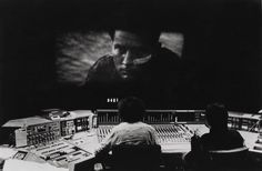 black and white photograph of two men sitting in front of sound mixing equipment with a large screen behind them