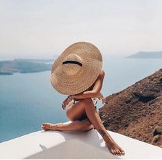 a woman wearing a straw hat sitting on the edge of a cliff looking at the ocean