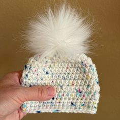 a hand holding up a white knitted hat with a pom - pom