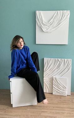 a woman sitting on top of a white chair next to two paintings and a wooden floor