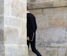 a man in black jacket and pants leaning against a stone wall with his foot on the ground