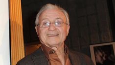 an older man wearing glasses and a brown shirt smiles at the camera while standing in front of a wooden column