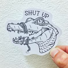 a hand holding up a sticker that says shut up with an alligator's head