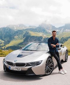 a man sitting on the hood of a silver sports car in front of some mountains