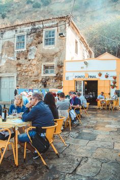 people are sitting at tables outside in front of an old building, eating and drinking