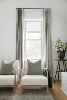 two white chairs sitting in front of a window next to a vase with flowers on it