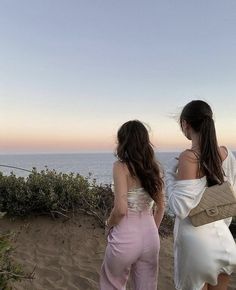 two young women standing on top of a sandy hill next to the ocean at sunset