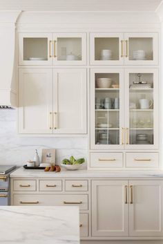 a kitchen with white cabinets and marble counter tops, gold pulls on the glass doors