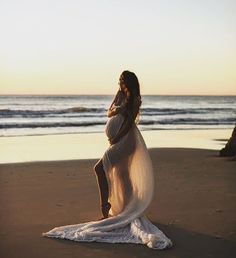 a pregnant woman standing on top of a sandy beach next to the ocean at sunset