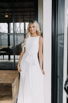 a woman standing in front of a window wearing a white dress with a bow at the waist