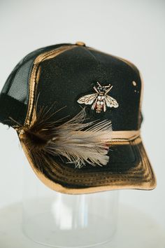 Look sharp and show off your style with this GOLDEN BEE BLACK TRUCKER HAT. This unique hat features a black trucker hat with gold accents, a gold bee patch, and a black and white feather combo that is sure to turn heads! Rock this hat and look cool and stylish - no bee-lieving required! This is a made-to-order item. All customized orders are currently shipping within 14 business days. To receive item quicker, expedited shipping is available at checkout. Bridal Trucker Hats, Cool Hat Designs, Women’s Trucker Hats, Hand Painted Trucker Hats, Diy Trucker Hat Ideas, Cute Trucker Hats, Painted Trucker Hats, Custom Hat Ideas, Trendy Hats For Women