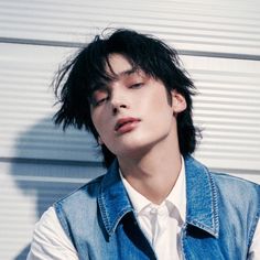 a young man with black hair wearing a denim vest and white shirt, leaning against a wall