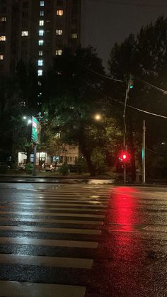 a red traffic light sitting on the side of a wet road at night with tall buildings in the background