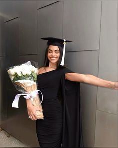 a woman in a graduation cap and gown holding flowers, standing next to a wall