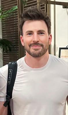 a man with a beard wearing a white t - shirt and carrying a black backpack