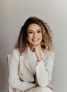 a woman with curly hair sitting on a chair smiling at the camera and wearing a white blazer