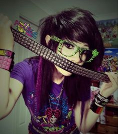 a girl with long hair wearing glasses and holding a guitar strap to her face while standing in front of a wall