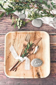 a wooden plate topped with silverware and greenery next to a couple of rocks