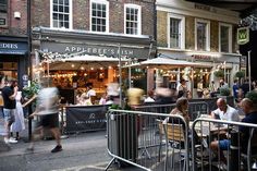 people sitting at tables in front of an outdoor restaurant on a city street with shops and restaurants