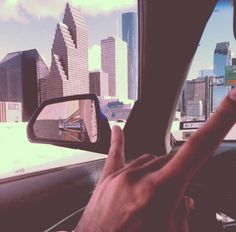 a person's hand on the steering wheel of a car in front of a cityscape