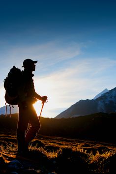 the silhouette of a man with a backpack walking through a field in front of mountains