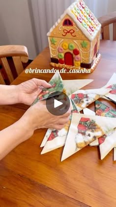 someone cutting out christmas cards on a table with a gingerbread house in the background