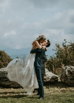 a bride and groom embracing each other in front of some rocks on the grass with mountains in the background