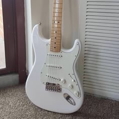 a white electric guitar sitting on top of a carpeted floor next to a window