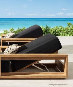 two wooden lounge chairs sitting on top of a cement floor next to the ocean and blue sky