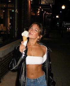 a woman is eating an ice cream cone on the street at night, wearing jeans and a leather jacket