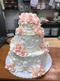 a three tiered white cake with pink flowers on it sitting on a wooden table