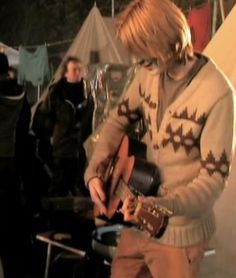 a man playing an electric guitar in front of other people at a camp site with tents