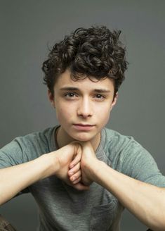 a young man with curly hair is posing for a photo in front of a gray background