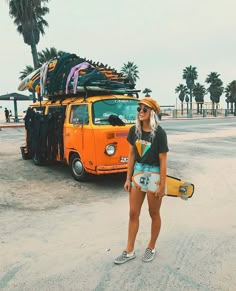 a woman holding a skateboard standing in front of an orange van with surfboards on top