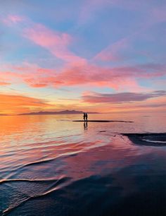 two people are standing on the beach as the sun goes down in the distance with pink and blue clouds