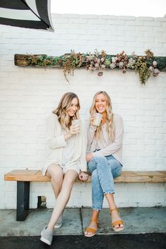 two women are sitting on a bench and drinking coffee in front of a white brick wall